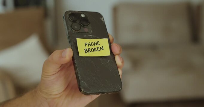 Person holding a damaged smartphone with a &ldquo;Phone Broken&rdquo; note attached.