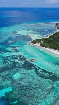 Drone shot of vast shallow coral reef lagoon leading to an inhabited tropical island with boats. Great background for travel, nature, and island lifestyle promo.Himmafushi.Maldives.vertical video