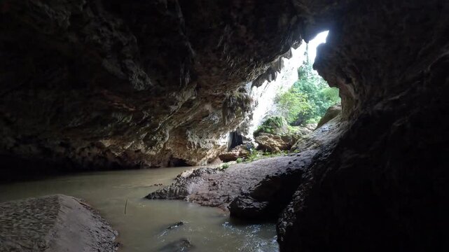 Dark Cave Entrance with Flowing River and Bright Green Jungle Outside in Thailand