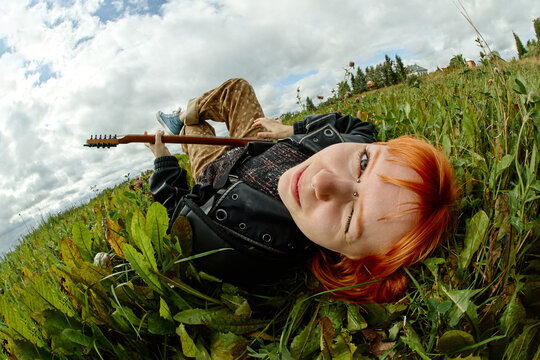 Portrait of Caucasian gen z young adult woman musician lying in green grass holding guitar, representing youth lifestyle and creativity. Image suited for indie music festival branding