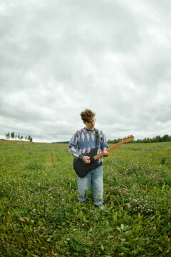 Vertical image of Caucasian young adult musician playing electric guitar in rural field. Visual for indie music production, creative youth lifestyle, and media advertising