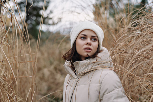 Woman in a beige winter coat standing among tall grass in an outdoor landscape, portrait style with calm mood and soft natural light.
