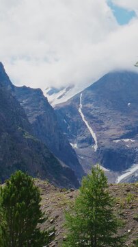 Stunning alpine valley with snow-capped peaks and glaciers. Larch trees stand tall against rocky slopes. Clouds drift around mountain summits. Scene evokes awe and adventure
