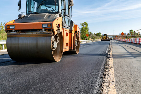 Orange road roller compacts fresh asphalt. Worker operates heavy machinery on construction site. Sunlight glints off metal and pavement. Road stretches ahead with safety barriers