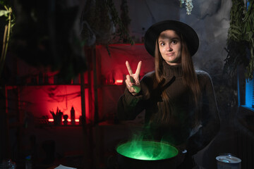 Realistic witch brewing a potion in cauldron showing victory V gesture. Blend of everyday life with traditional witchcraft, magic and mystical atmosphere © Andrii Lysenko