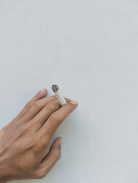 Close up of a human hand holding a traditional lit hand-rolled tobacco cigarette against a plain white wall background