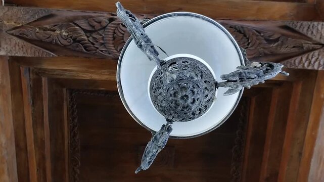 Antique Javanese hanging oil lamp modified with electric bulb under carved teak Joglo roof ceiling, vintage traditional interior lighting