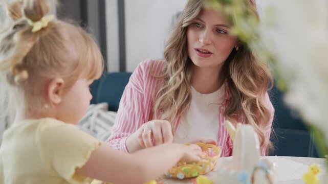 Young girl and mother interact warmly at table. Girl holds colorful cupcake, mother smiles gently. Scene captures joyful, tender moment of shared activity