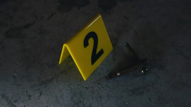 Close up of a yellow evidence marker with the number two next to high caliber bullets on a dark, dusty concrete floor, representing a crime scene investigation or forensic analysis