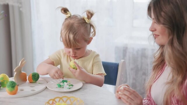 Young girl decorates Easter egg with green paint. Mother watches with gentle smile nearby. Soft morning light fills room with warmth. Child focused, concentrated on creative task