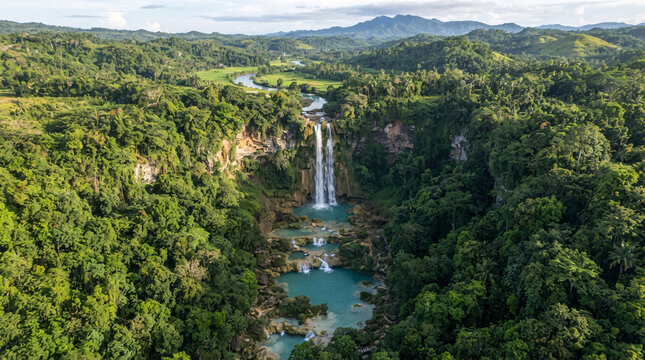Aerial panoramic view of a spectacular tropical waterfall plunging into turquoise pools within a vast lush rainforest valley surrounded by rolling green hills and distant mountains.