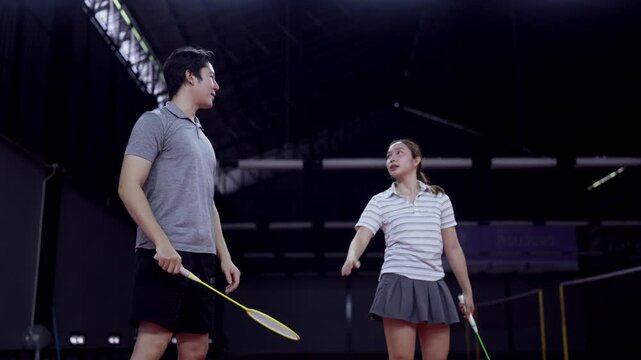 Asian male coach guiding young girl through badminton swing technique on indoor court