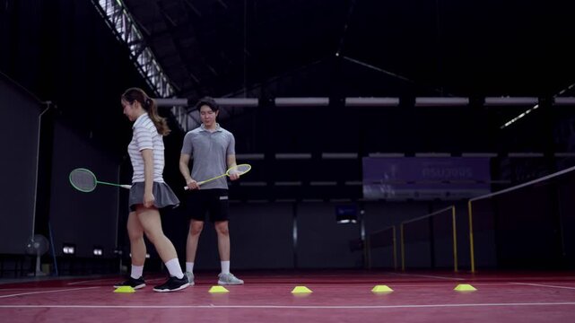 Male badminton coach guiding young woman through footwork drill using training cones on indoor court