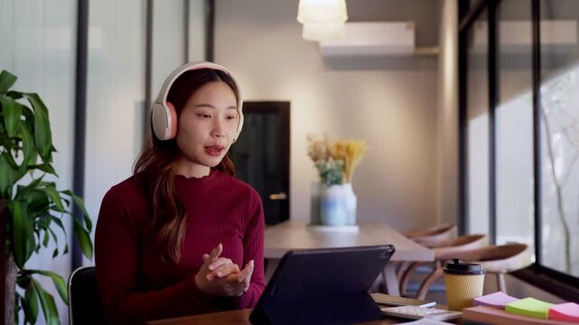 Young Asian woman wearing headphones gestures during video call on tablet in modern office