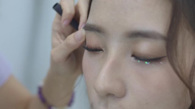 Extreme close up of a makeup artist applying eyeliner and glitter eye makeup to the eyes of a young Asian woman with false lashes filmed in 4K