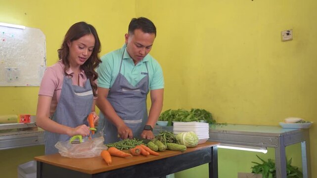 Adults in aprons peel carrots and cut green beans at metal kitchen worktable, arranging plates and bowls in bright yellow domestic cooking scene