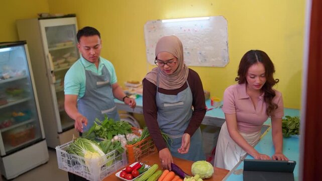 Three young adults handle fresh produce and tablet payment inside compact neighborhood shop, with one man holding cabbage, woman in hijab sorting goods.