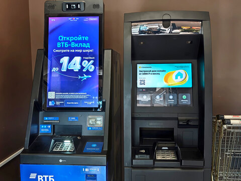 Kirov, Russia - June 17, 2026: VTB bank self-service terminal and ATM stand side by side indoors with digital screens showing financial ads