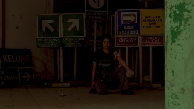 Wide shot of a wounded female hostage sitting alone in front of traffic signs in a dark, creepy underground structure, looking exhausted and scared.