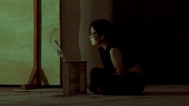 Side angle of a woman sitting on the ground in a gloomy, dilapidated room, her face illuminated by her computer. Concept of a secret underground cyber operation.