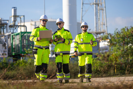 Team of engineers working with laptop and tablet at industrial plant, diverse manufacturing team walking together with safety equipment near refinery refinery factory under blue sky