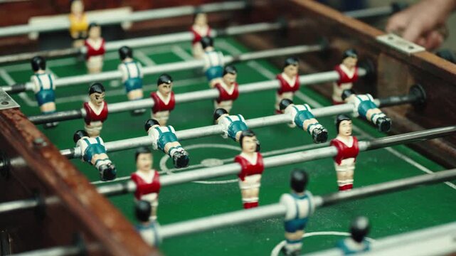 Red and blue striped figures lined up on the rods of a foosball table, with a white ball in play and a hand on a handle.