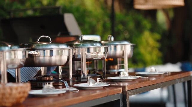 A row of chafing dishes with lids and a bread basket on a wooden buffet table outdoors, with plates set for guests.