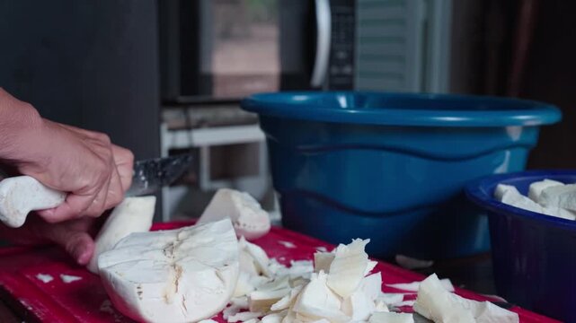 Female hands using a knife to peel and chop fresh cassava root on a red cutting board. Rural food preparation for preservation at home