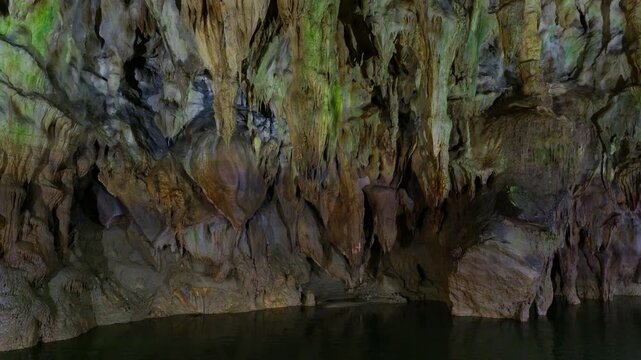 Underground cave river flows through limestone chamber with hanging stalactites and mineral-stained rock formations. Static wide angle shot captures the natural geological features and dark water.