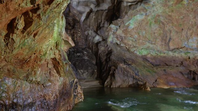 Interior view of Aggitis Cave reveals an underground river with green-tinted water flowing between orange and brown mineral-stained rock walls. Smooth upward tilt shot reveals layered geological.