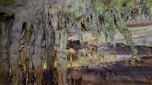 Stalactites and mineral formations hang above an underground lake inside Aggitis Cave, reflecting in the still water. The shot captures the layered rock ceiling with moss and mineral deposits.