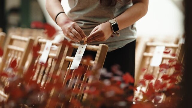 A woman ties paper name tags onto the backs of wooden chairs before a wedding, with red leaves in the foreground.