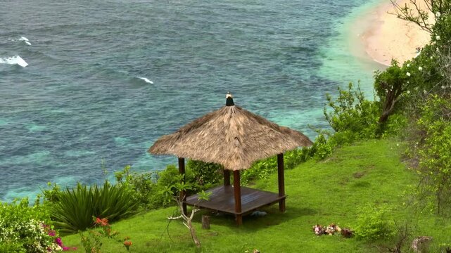Straw roof gazebo providing a serene viewpoint over clear blue water, a small sandy beach, and lush green vegetation on a tropical island cliff, offering a peaceful escape on tropical Bali, Indonesia