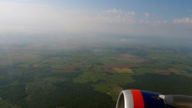 Airplane window view of green farmland, forests and villages below, jet engine nacelle in frame, hazy summer sky, sun flare