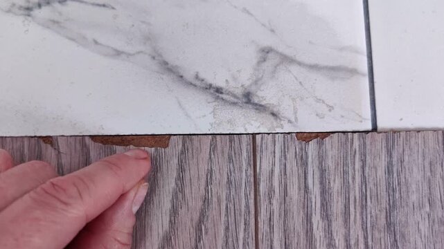 A hand picks at a chipped edge where tile meets laminate flooring, exposing a damaged surface, home repair problem, renovation and maintenance detail.