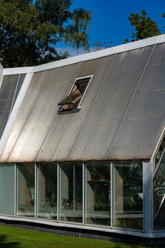 Open roof window in a modern greenhouse. Ventilation hatch on a sloped polycarbonate roof with glass walls and green trees. Garden architecture.