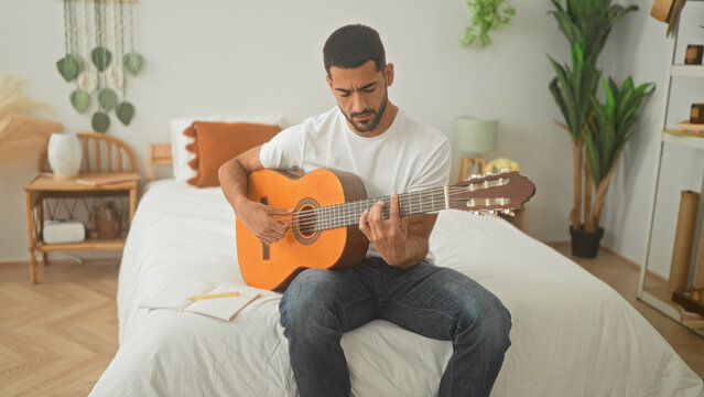 Man playing an acoustic guitar on a bed with hands fretting strings in a studio space, jotting lyrics in a notebook beside him; songwriting calm.