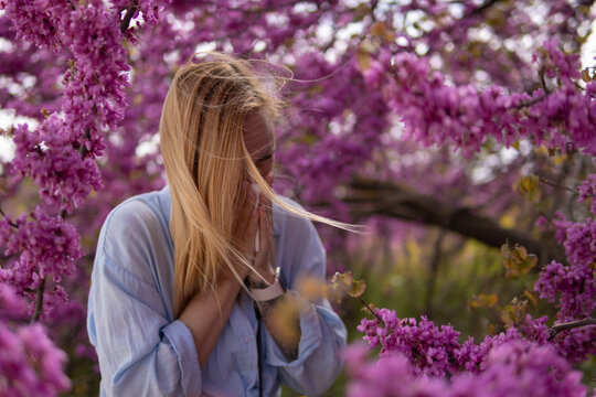 Young woman experiencing severe pollen allergy symptoms and hay fever, sneezing and rubbing her nose amidst vibrant purple flowers blooming in a spring garden outdoors