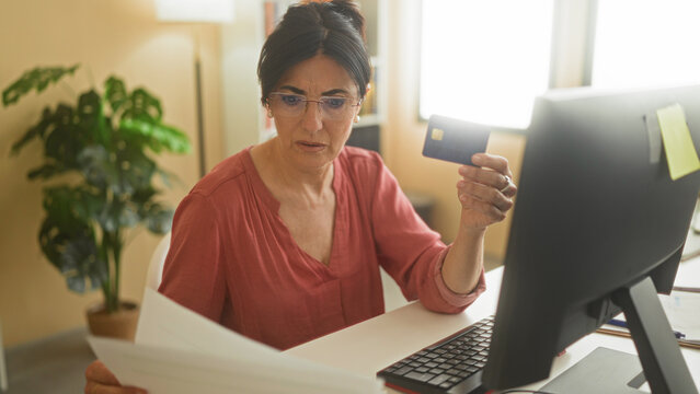 Woman holding creditcard and reading paperwork at desk in a building, looking concerned at computer screen; financial concern.