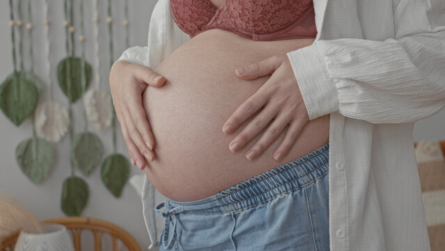 Pregnant woman cradling bare belly with both hands in studio near macrame wall and rattan chair, wearing open shirt and lace bra; serene anticipation.