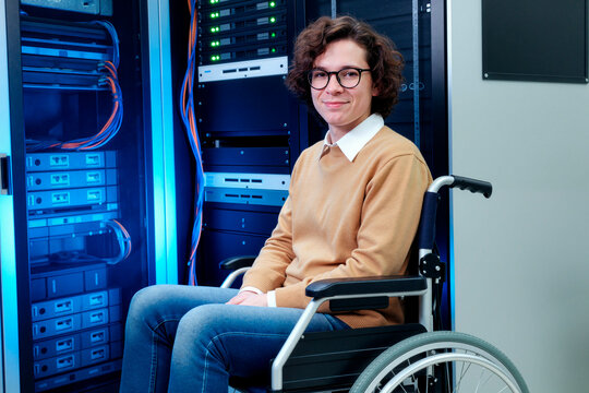 Portrait of Caucasian young adult man with disability sitting in wheelchair near server racks, working in IT infrastructure setting. Ideal for inclusion, tech careers, accessibility marketing