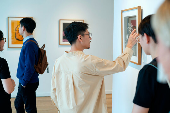 Asian young adults viewing framed artwork in gallery, discussing exhibition and pointing at details. Ideal for culture marketing, creative events promotion, museum campaigns
