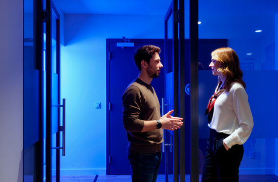 Caucasian young adult man and woman discussing project in office corridor, standing face to face and gesturing during meeting. Ideal for teamwork, corporate communication, partnership