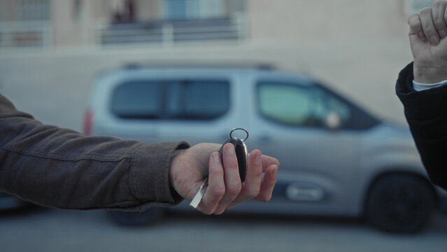 Young hispanic man receives car keys with open palm while another hand drops the fob beside a parked car on street; trust transfer.