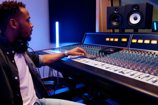 Young Black man adjusting audio mixing console in recording studio, monitoring sound production and music editing. Ideal for content about music industry, audio engineering, media production