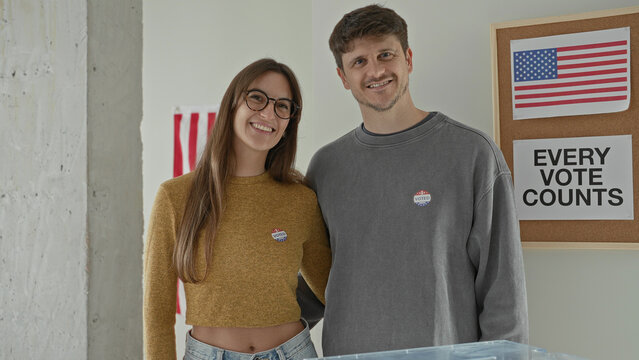 Young woman points finger to voted sticker on chest beside partner at ballot box in polling station building; civic engagement pride.