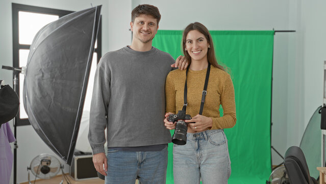 Young man and woman photographers in studio, woman holding camera and man hand on her shoulder beside green screen and softbox; creative collaboration joy.