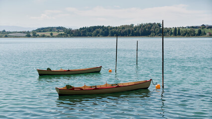 Pair of traditional wooden rowing boats moored to wooden stakes with orange floating buoys on clear turquoise lake water by a green forested coastline under sunny skies. © Ekaterina