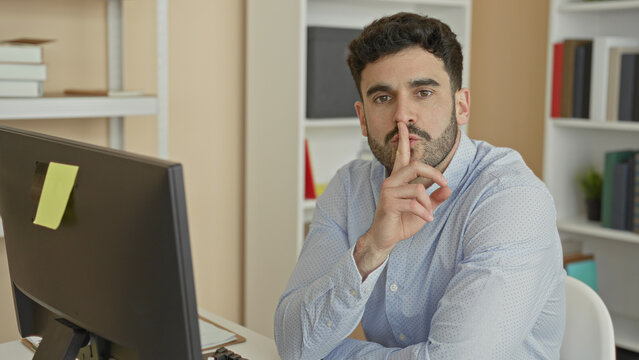 Man at computer with finger to lips performing shushing gesture in office building; quiet concentration.