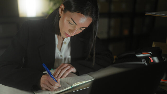 Woman writing in a notebook with laptop and barcode scanner on a desk in an office building at night; concentration diligence.